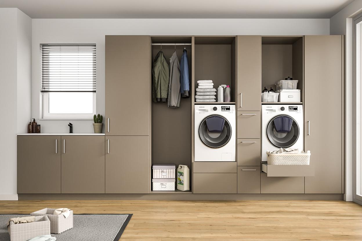 Ein heller Raum mit einem großen, maßgeschneiderten Schranksystem in Taupe, integrierten Waschmaschinen, Ablageflächen und einem Fenster mit Jalousien.